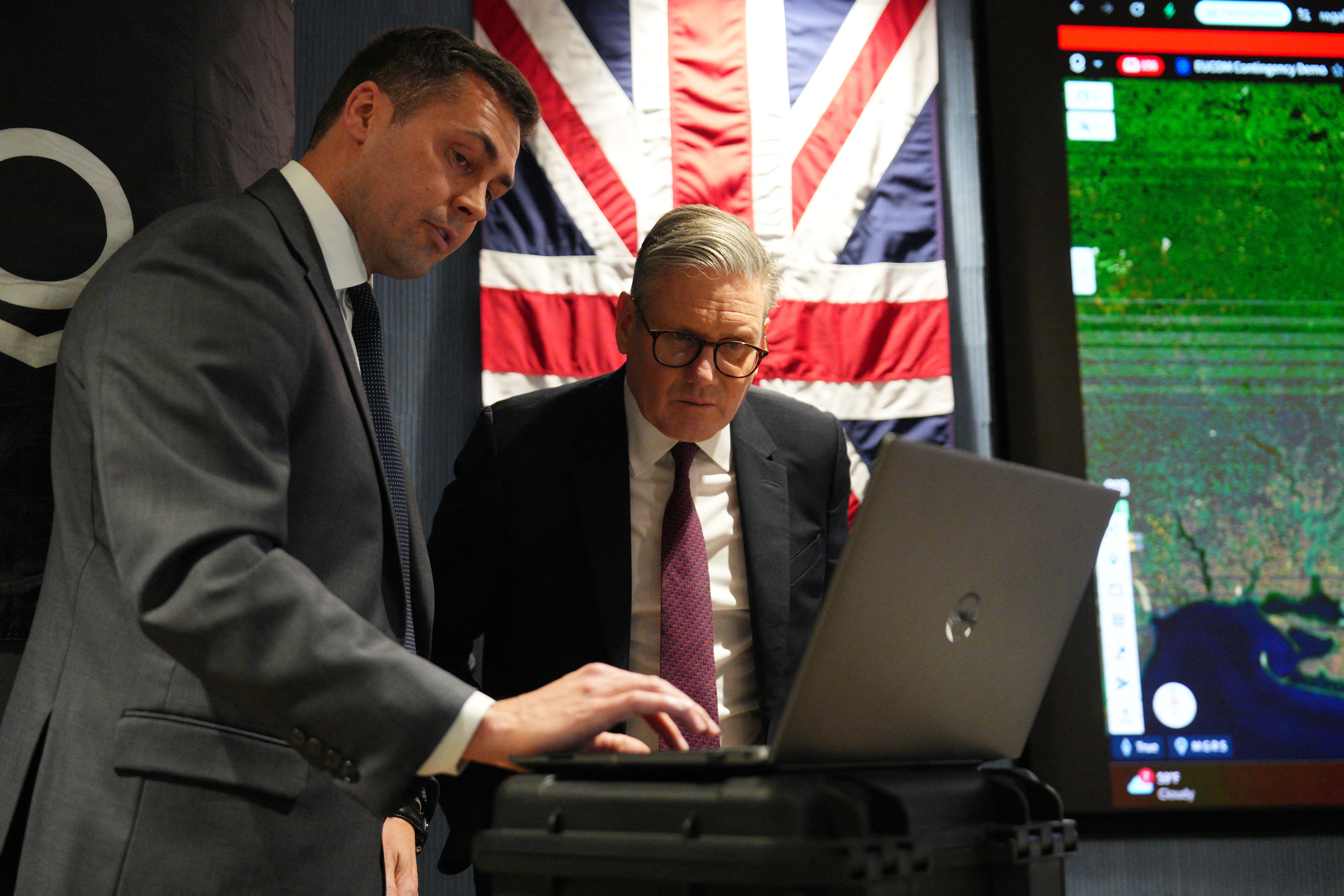 Keir Starmer and another man look at a laptop and screen with a map of the world.