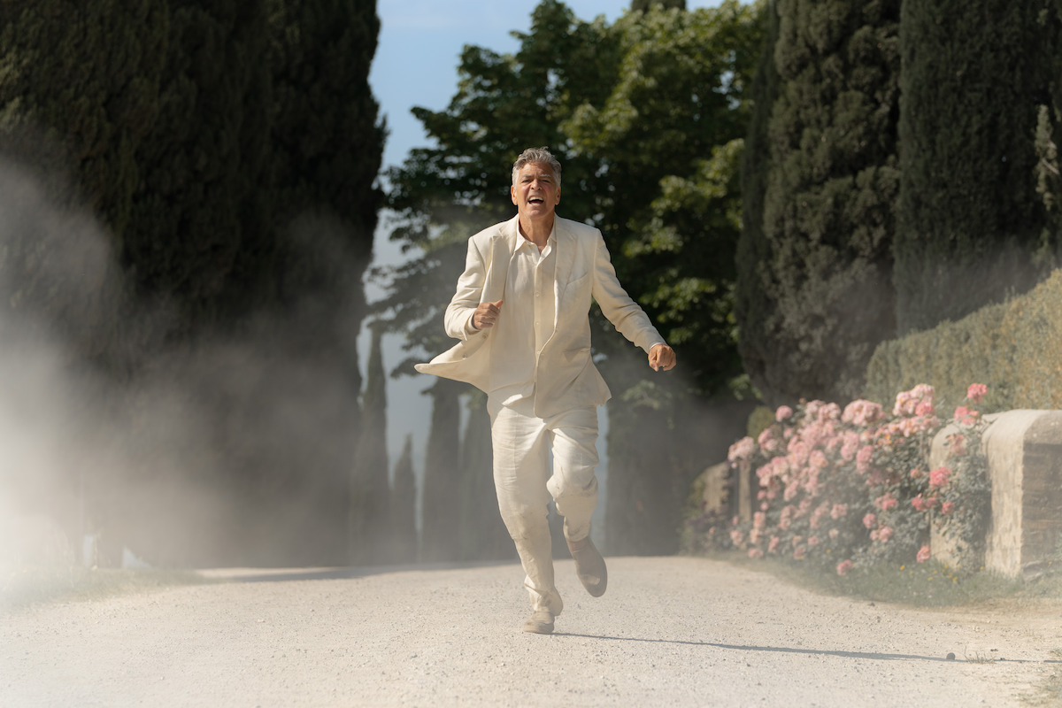 George Clooney, dressed in a light-colored suit is running down a tree-lined, dusty path with pink flowers on one side.