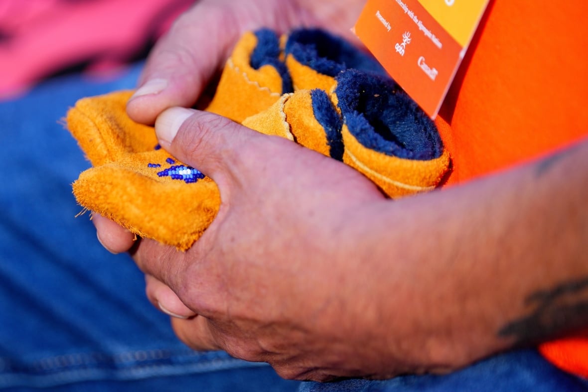 Someone holds a small pair of moccasins in their lap. The moccasins have a blue and white beaded pattern on them.