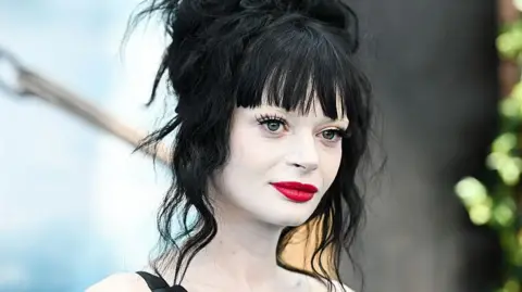 Getty Images Ruth Codd has pale, white make-up covering her neck and shoulders. She has bright red lipstick and dark mascara. She is smiling slightly, looking vaguely towards the camera, with a light blue and black backdrop at a press event for a recent film release.