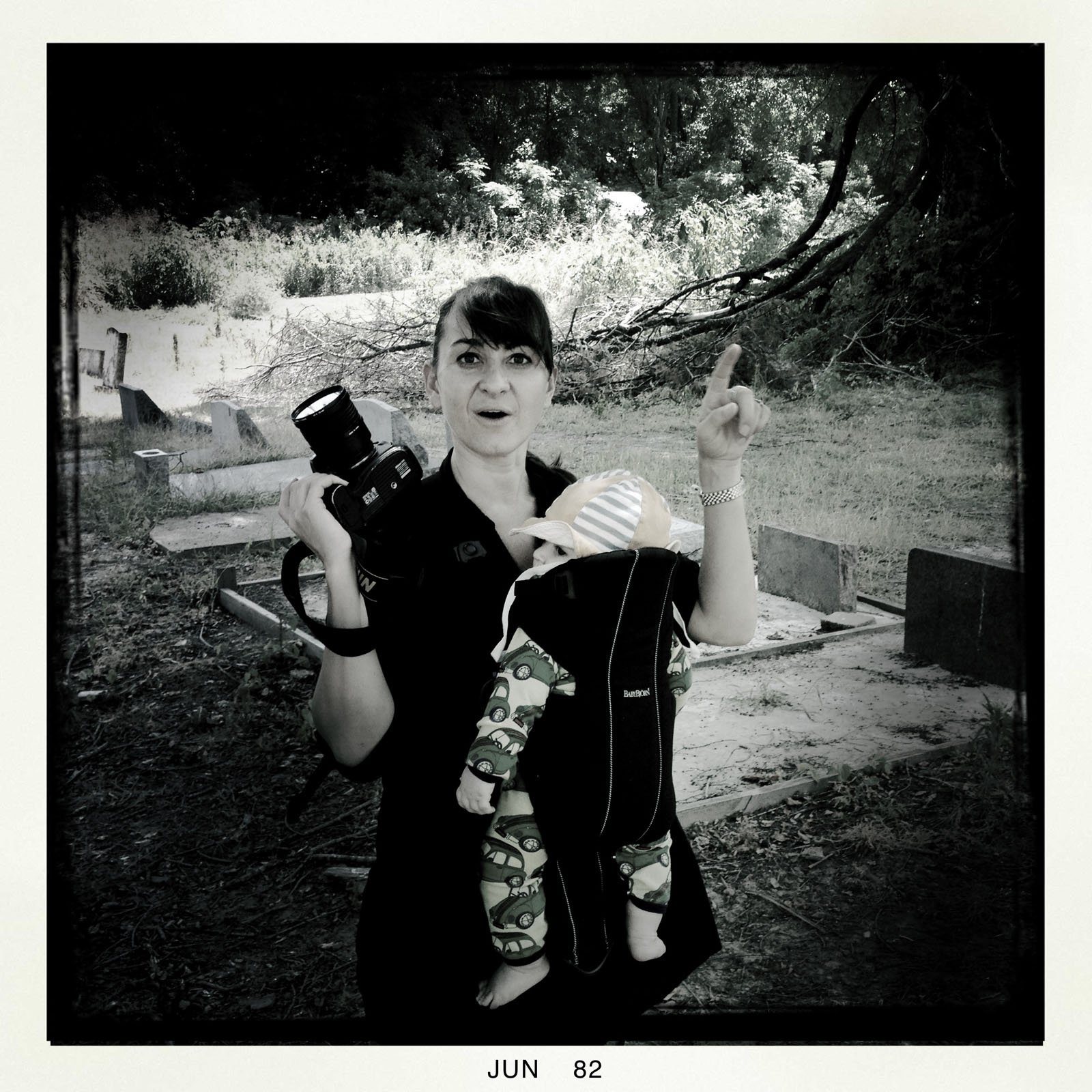 A woman holding a camera and wearing a baby in a front carrier stands outdoors in a grassy, wooded area. She looks surprised and points upward with one finger. The image is in black and white.