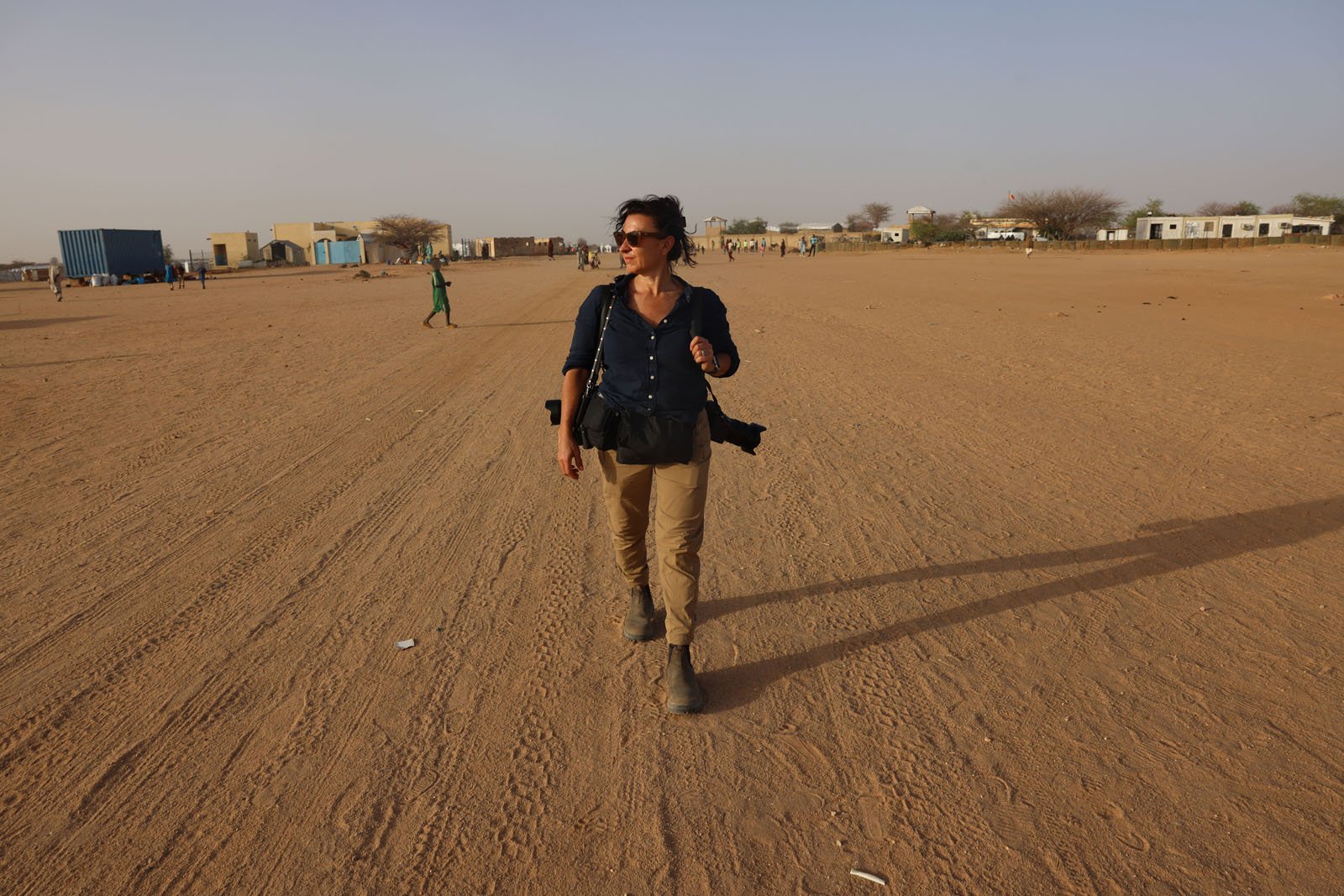 A woman wearing sunglasses, a dark shirt, and khaki pants walks alone on a vast, sandy area with scattered buildings and a few people in the distant background under a clear sky.
