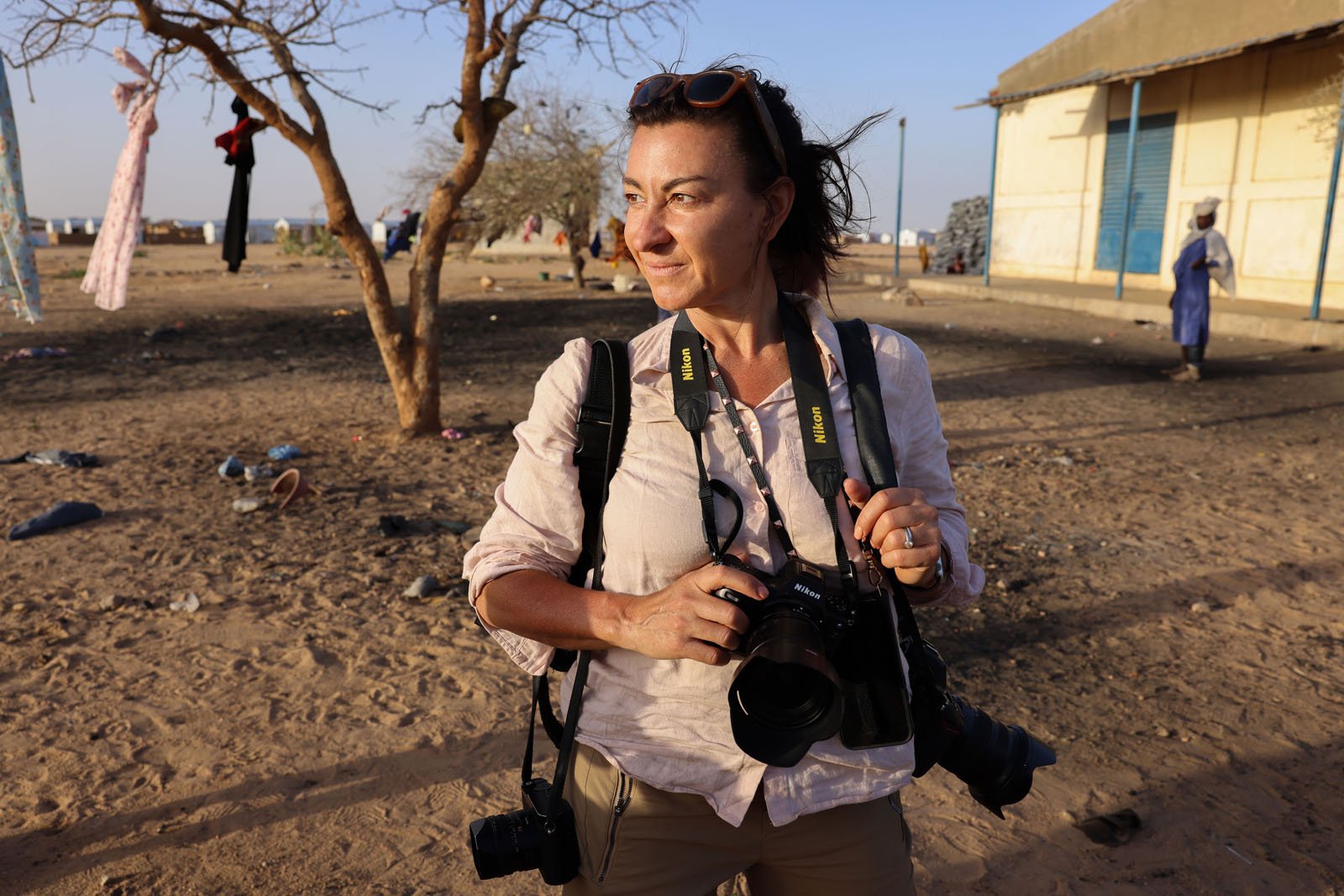A woman with dark hair, wearing a light shirt and sunglasses on her head, stands outdoors holding two cameras. She is looking to the side, with a dry, sandy landscape and a tree in the background.