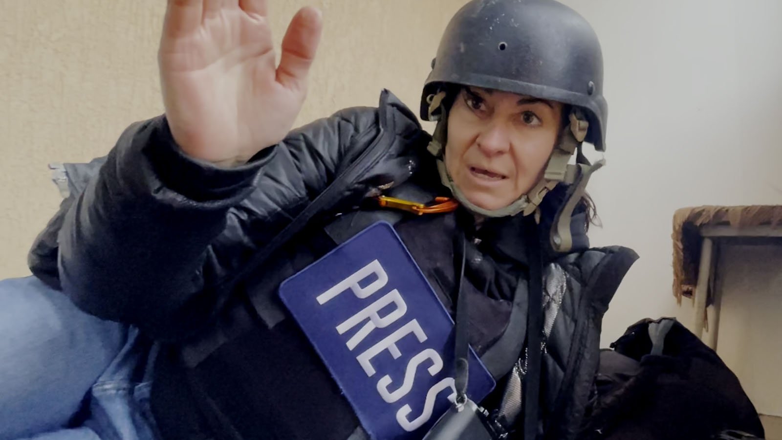 A journalist wearing a "PRESS" vest and a helmet sits on the floor indoors, raising one hand and looking tense, possibly in a conflict or dangerous situation.