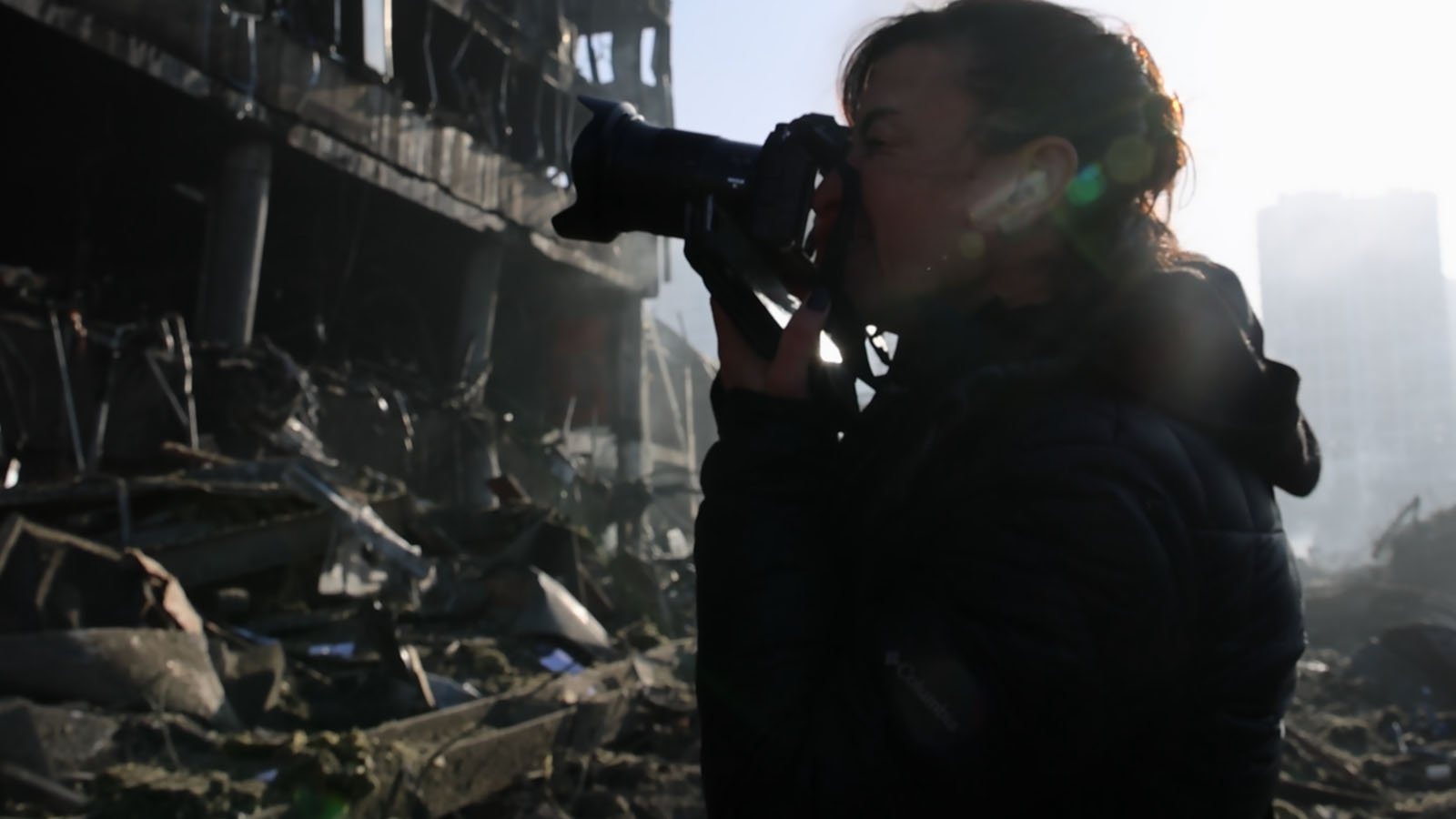 A person in a dark jacket takes photos with a camera amid rubble and debris in front of a damaged building, with sunlight shining from behind.
