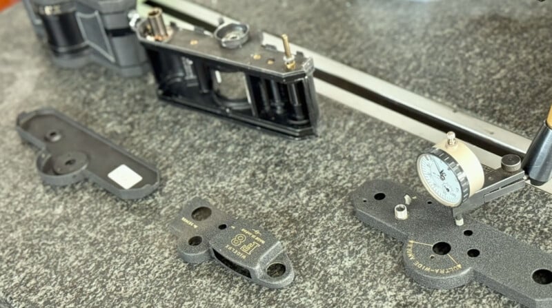 Various metal mechanical parts and measuring instruments, including a dial indicator, are arranged on a speckled granite surface, suggesting a precision measurement or calibration setup.
