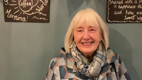 Liz sits in a cafe, she has shoulder-length hair and a fringe. She wears a blue and grey scarf and a patterned jacket