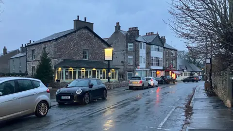 A photo of Silverdale in Lancashire. The picture shows some houses and the outside of a pub and some cars on the road.