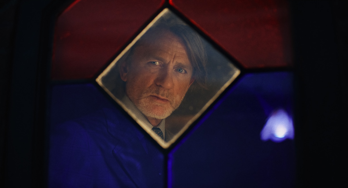 A serious middle-aged man in a suit looks through a stained glass window with red, blue, and clear panels, creating a contemplative and somber mood.