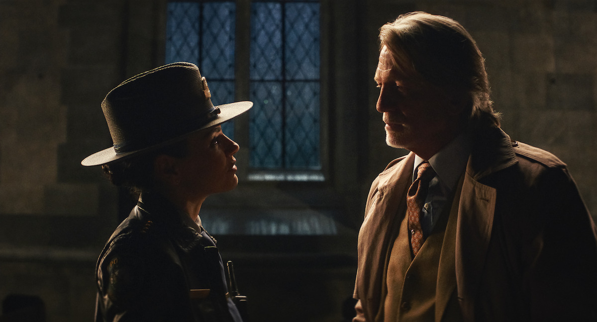 A woman in a police uniform faces an older man in a suit inside a dimly lit stone room with a large window; the mood is tense and serious.