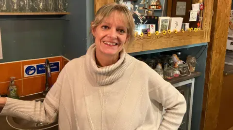 Nikki stands in the kitchen area of a coffee shop. A sink can be seen behind her with glasses on shelves above. She has blonde hair pulled away from her face, she wears a cream oversized jumper. She smiles at the camera.
