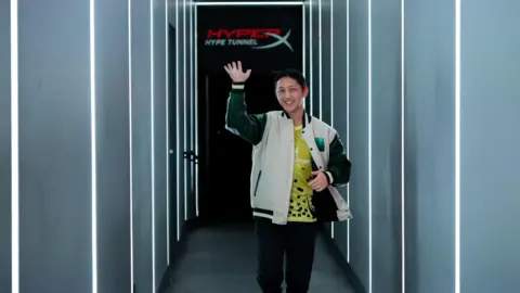 MEWC A man with short black hair emerges from "the HyperX Hype tunnel," his arm raised to wave ahead as he smiles. The tunnel is grey, with white light glowing through in large lines along the side and roof. The man is wearing a cream varsity jacket, underneath a yellow esports jersey with sponsors along it.