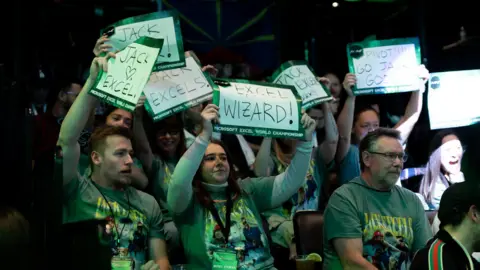 MEWC A crowd of people holding up signs with Excel related messages on them showing support for their favourite participant. One reads "Excel Wizard", and another says "Jack Excels."
They are all covered by a green light, which sweeps across the crowd and arena.