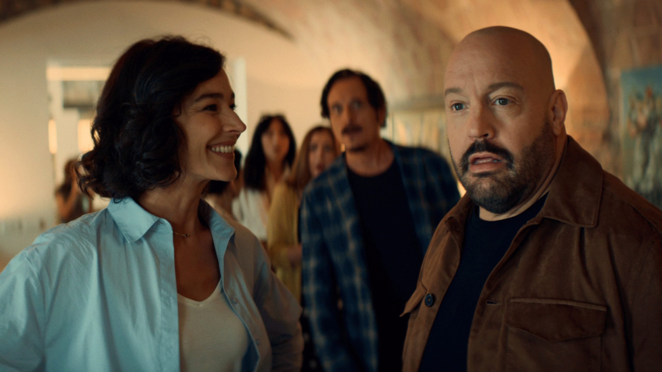 A group of adults stands indoors, with a woman in a light blue shirt smiling and a man in a brown jacket looking surprised in the foreground.