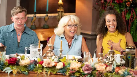 A man sat looking down at a table, sat next to two women laughing