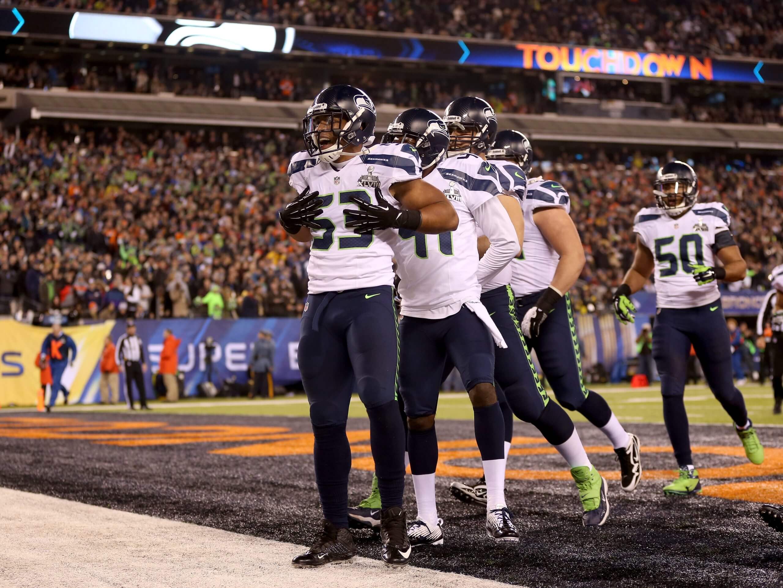 Malcolm Smith puts his hands to his chest and looks outward. He wears a white jersey with navy pants, and his teammates are behind him.