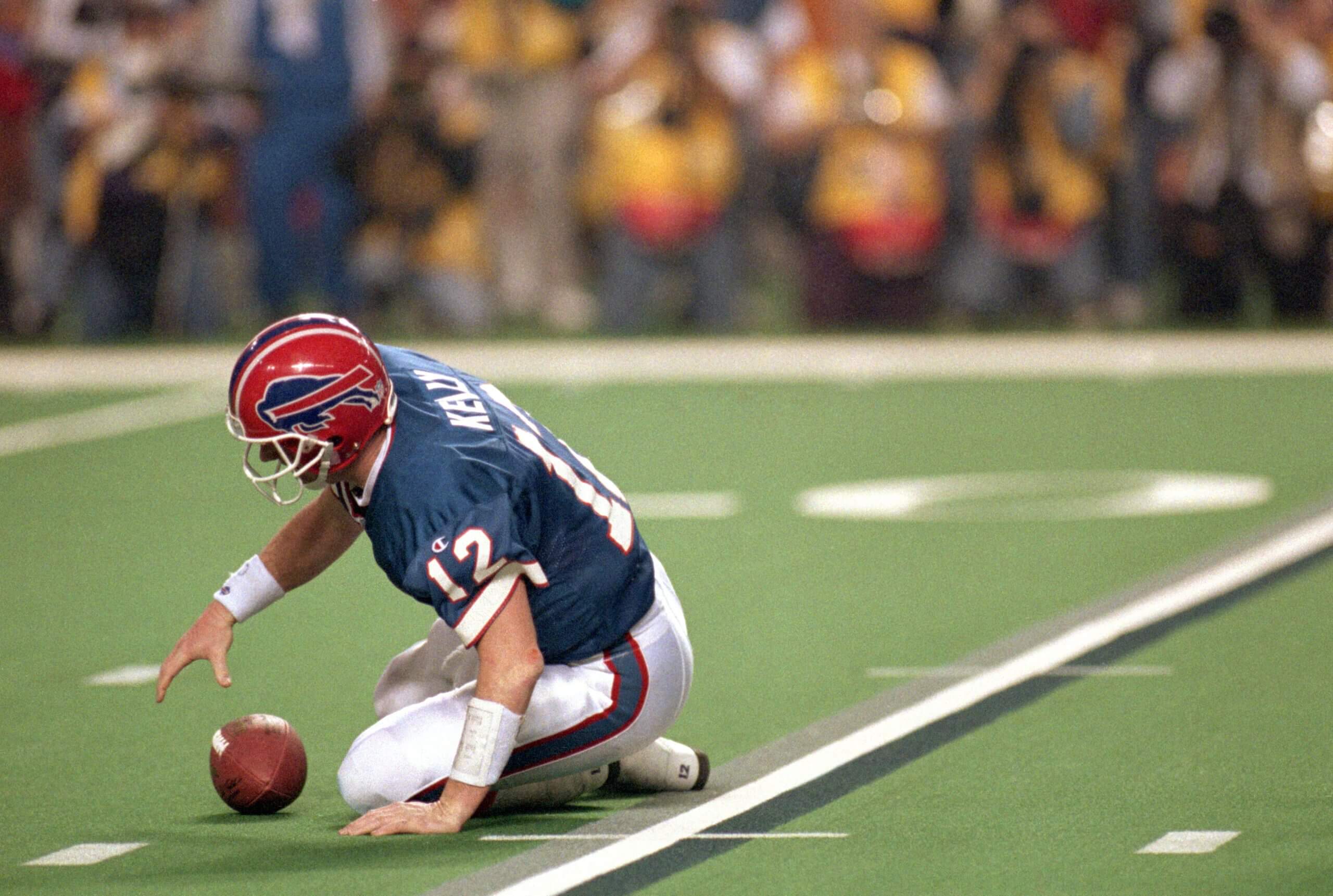 Jim Kelly reaches for the ball on the ground with his right hand. He's in a blue jersey and red helmet.