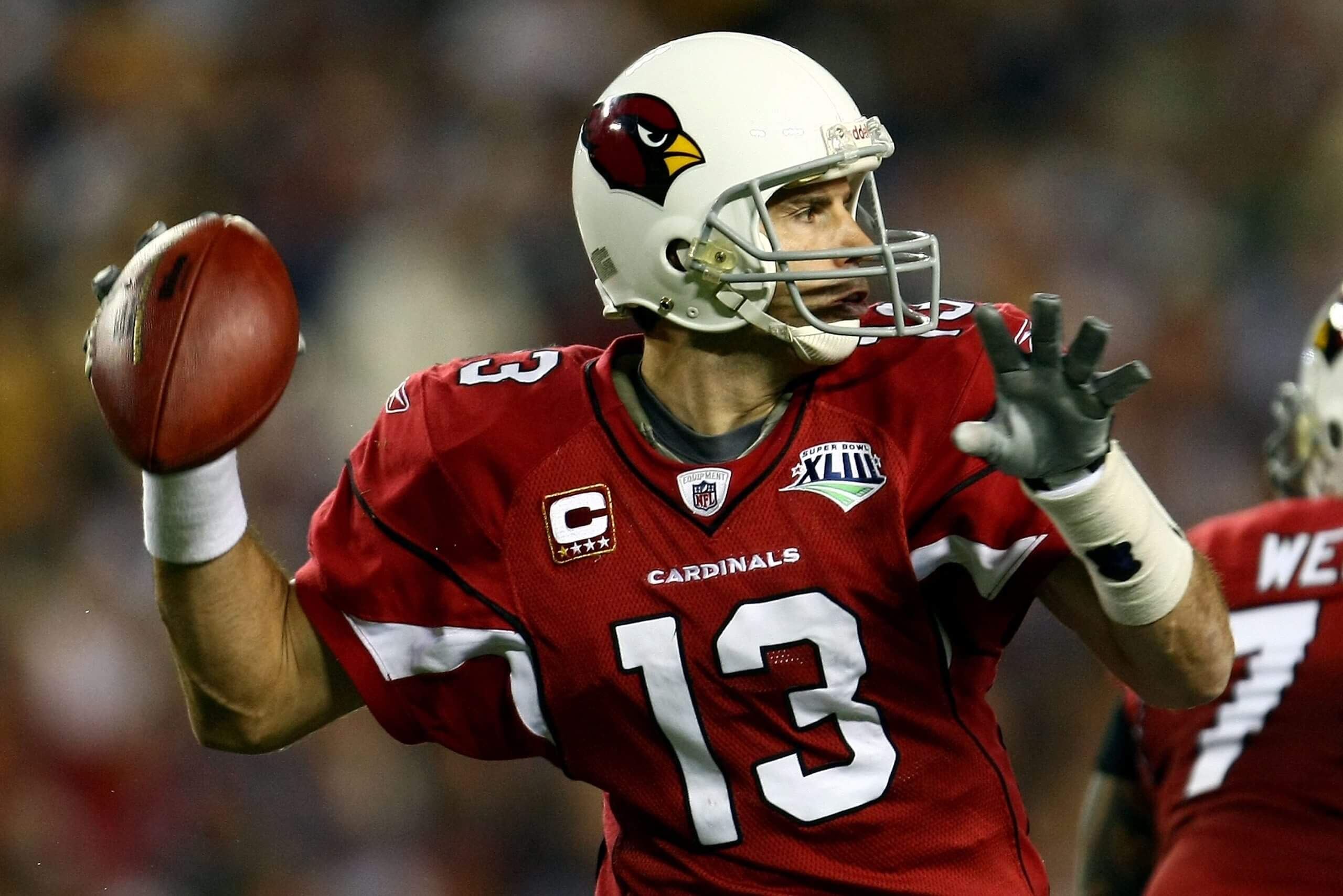Kurt Warner, in a red jersey and white helmet, winds up to throw a pass.