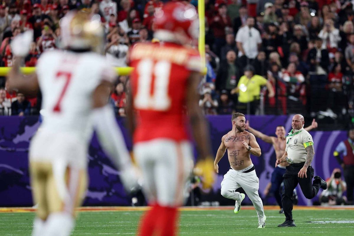 In an intense moment during an American football game, two players in distinct uniforms engage in play. One player, identifiable by the number 7 on his jersey, is seen sprinting across the field, while another player, wearing the number 11, follows closely behind. The referee is present at the scene, overseeing the action. The backdrop features a lively crowd, adding to the game's atmosphere.