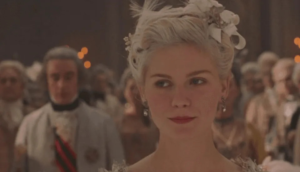 A woman in elegant historical attire at a formal gathering, with guests in the background. | IMDB