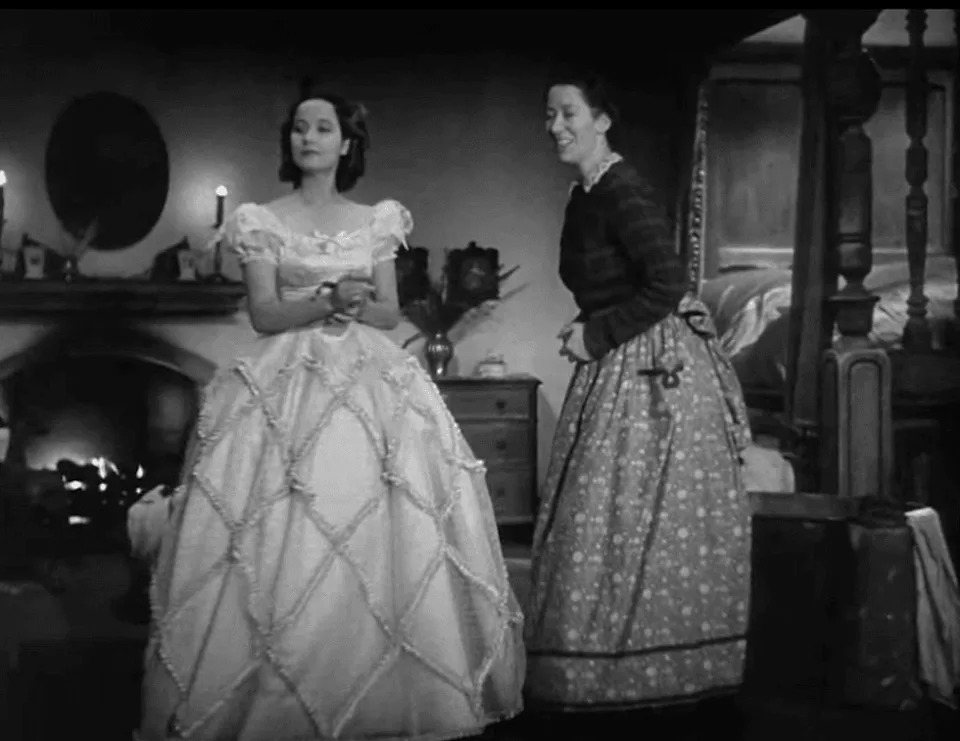 Two women in period dresses standing in a vintage room with a lit fireplace. | IMDB