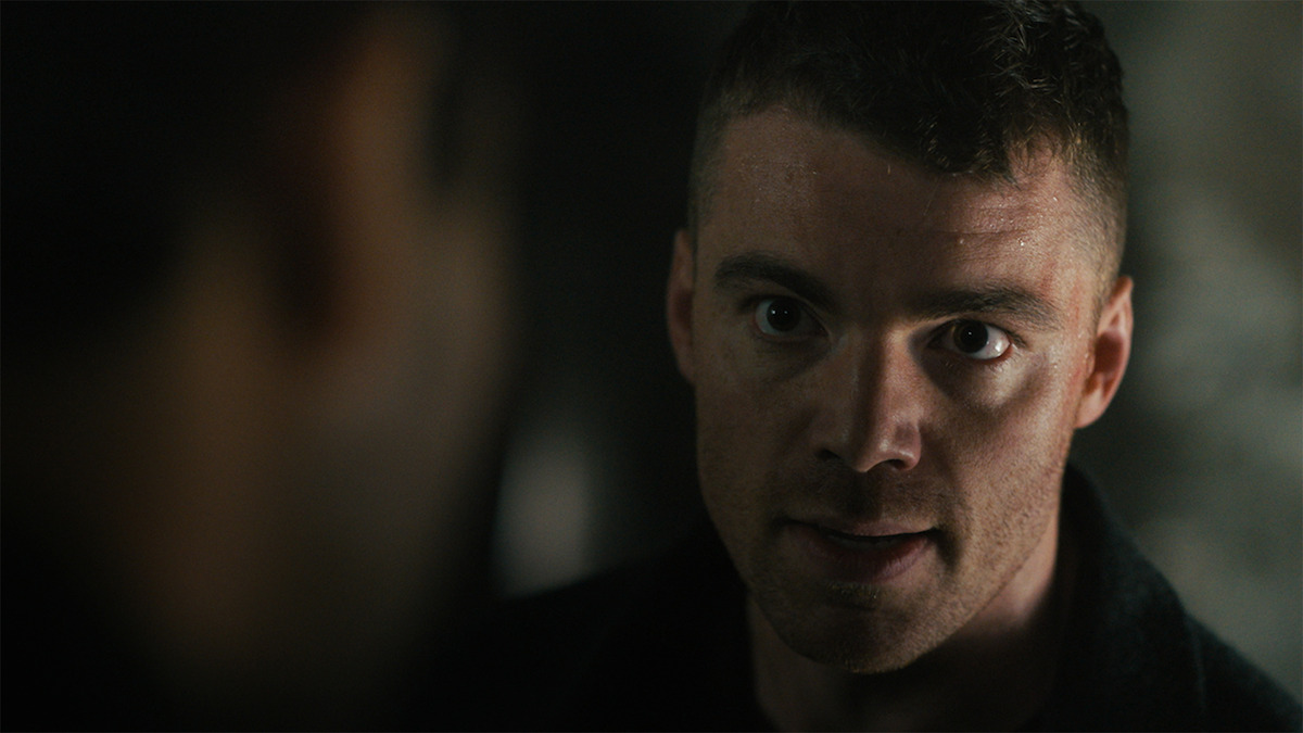 A man with a tense, intense expression confronts another person in a dimly lit, shadowy environment, highlighting dramatic tension.