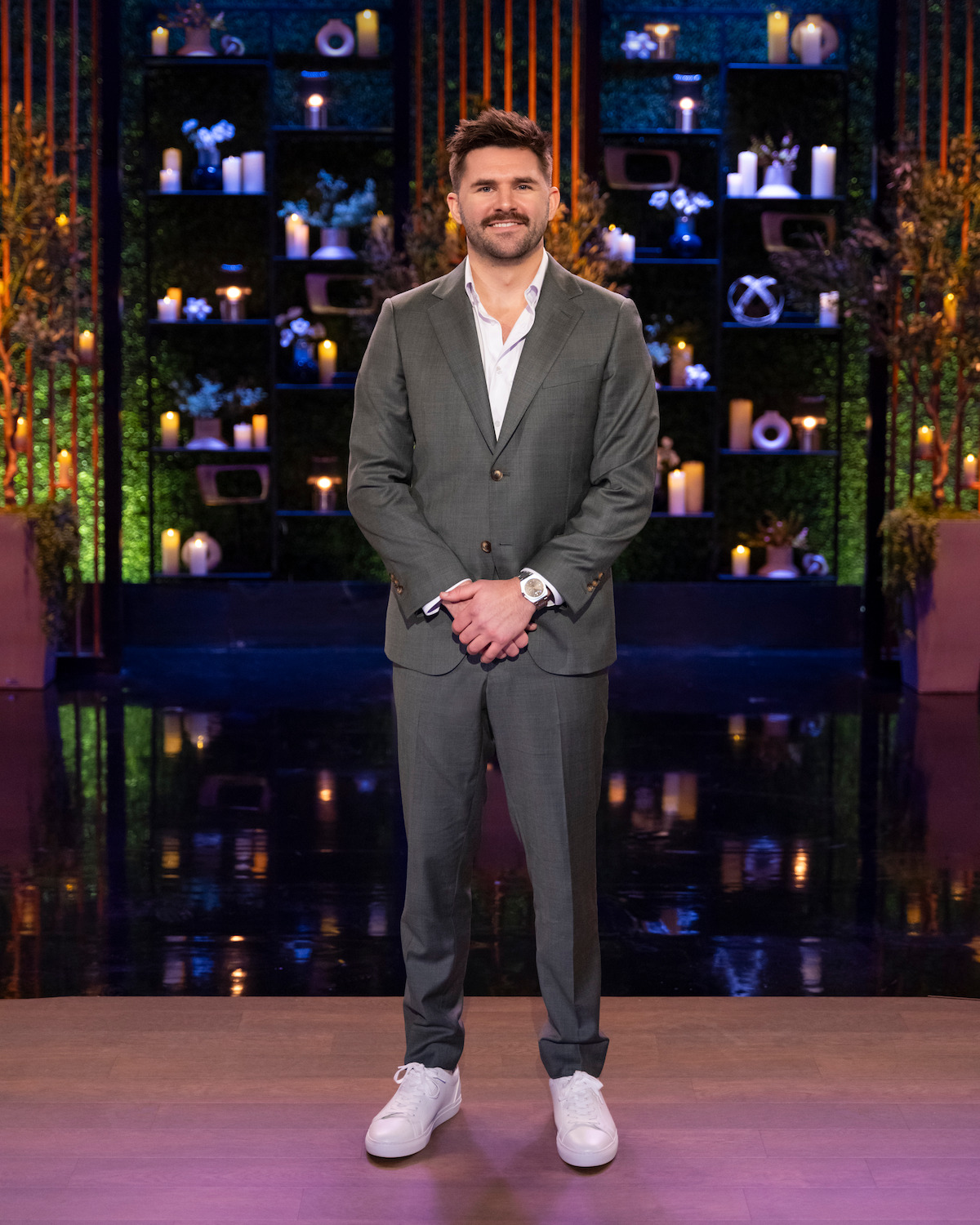 Man in a gray suit and white sneakers stands on a stage with candles, plants, and decorative shelves in the background, under studio lighting.