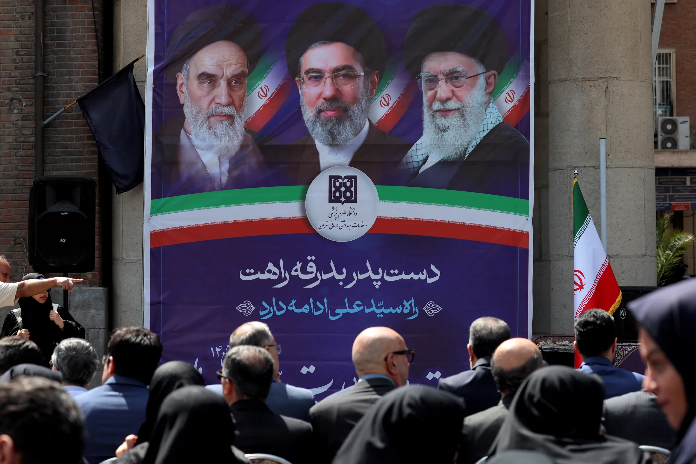 People attend a rally against attacks on health facilities, sitting in front of a banner depicting three Iranian supreme leaders.