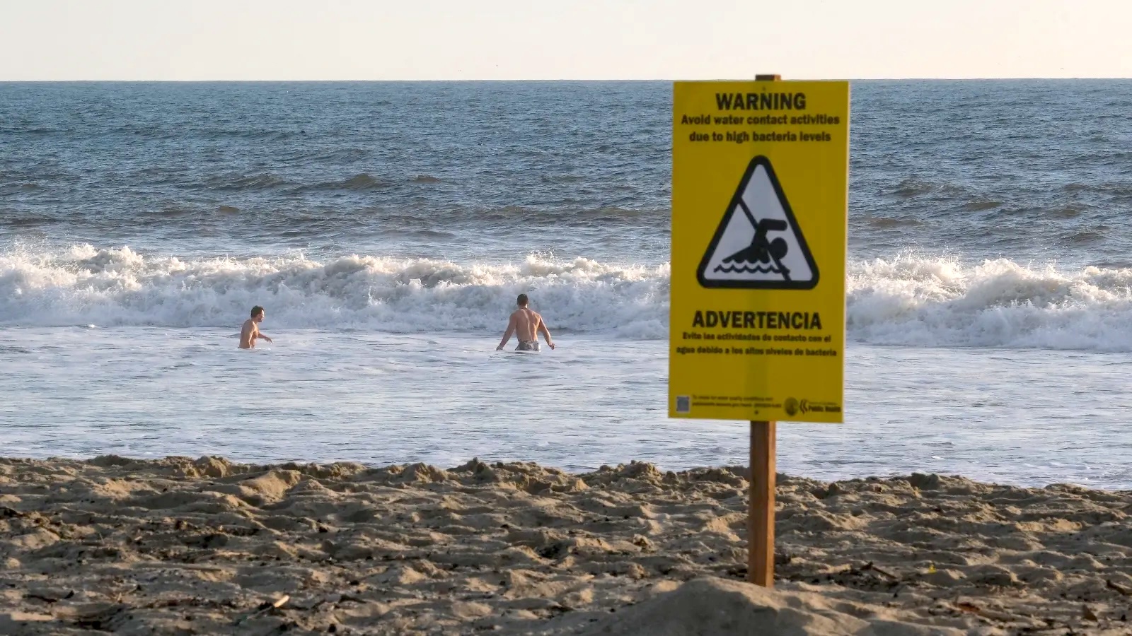 A sign on a beach warns of high bacteria levels while people swim in the water